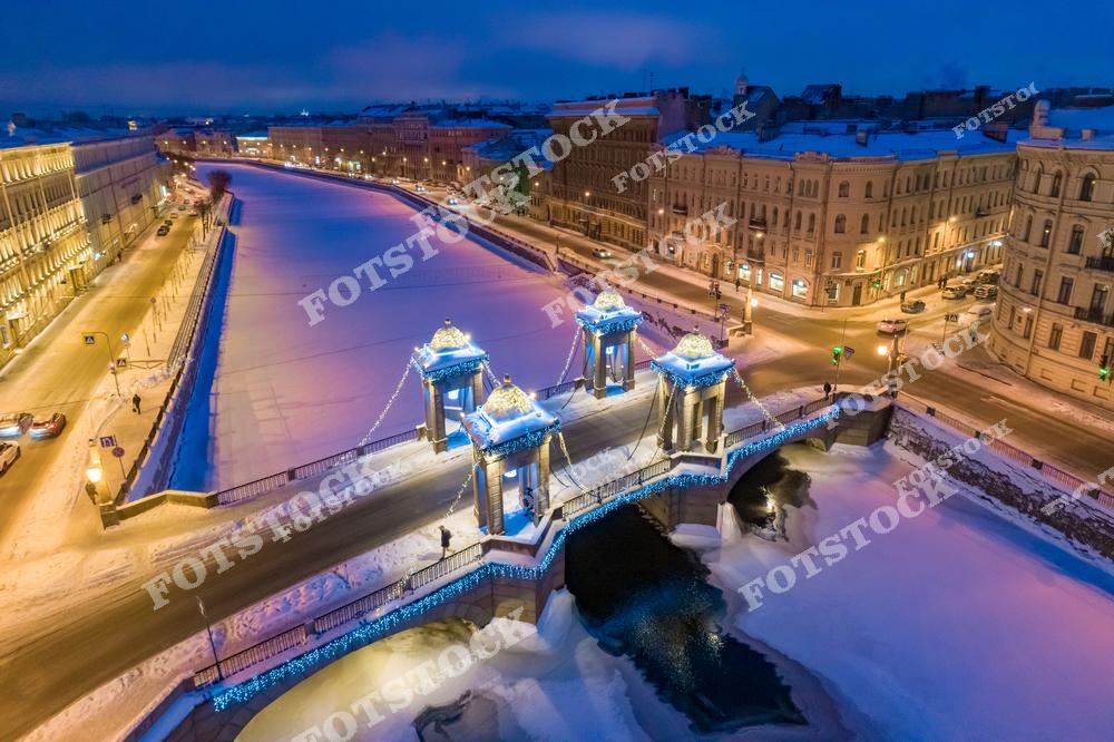 На фотографии изображен мост Ломоносова, один из старейших мостов Санкт-Петербурга, перекинутый через замерзшую реку Фонтанку. Вечерняя иллюминация подчеркивает его архитектурные детали, создавая волшебную атмосферу зимнего города. Окружающие исторические здания и заснеженные улицы добавляют картине уюта и спокойствия. Мост и замерзшая река освещены огнями, которые отражаются в ледяной глади.